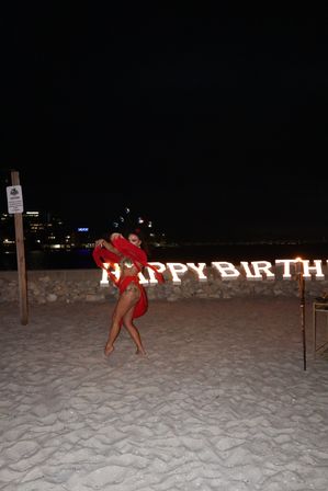 Dancer in a red costume performing on a sandy beach at night in front of illuminated HAPPY BIRTHDAY letters, with torches and distant city lights in the background.