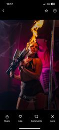 Nightclub performer in black costume wielding a handheld flame-thrower, shooting a tall flame beside a chrome pole under neon pink and purple stage lighting.