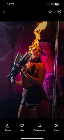 Nightclub performer in black costume wielding a handheld flame-thrower, shooting a tall flame beside a chrome pole under neon pink and purple stage lighting.