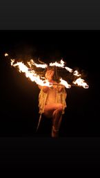 Fire dancer performing with a multi-wick flaming hoop above a crouched figure in a gold costume against a dark background