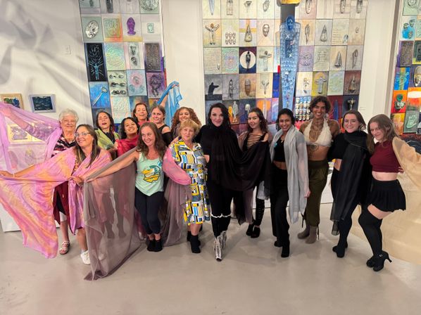 Group of women posing indoors in an art gallery in front of a colorful tiled mosaic wall, wearing eclectic outfits and flowing translucent scarves for a joyful community art event.