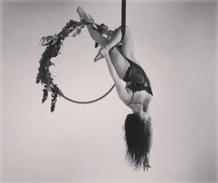 Black-and-white studio photo of an aerialist hanging upside down from a flower-wrapped lyra (aerial hoop), wearing a lace costume with hair cascading — graceful aerial performance.