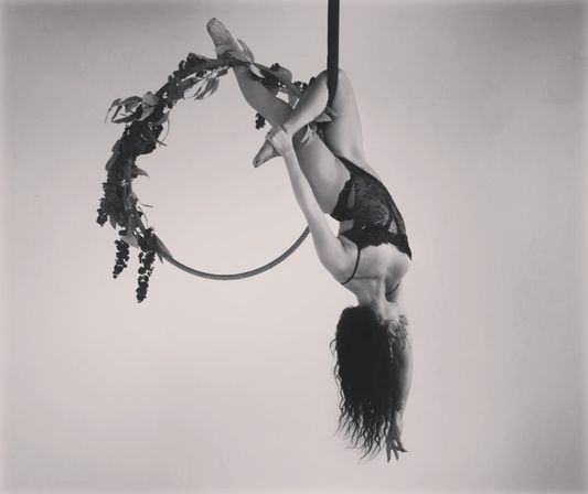 Black-and-white studio photo of an aerialist hanging upside down from a flower-wrapped lyra (aerial hoop), wearing a lace costume with hair cascading — graceful aerial performance.