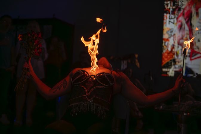 Burlesque fire performer leans back as a dramatic flame rises from their throat and chest, wearing a beaded corset and holding red roses and a lit torch on a dark club stage.