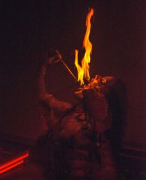 Close-up of a fire-eater performing a tall orange flame trick against a dark, dramatic background — night performance with glowing, dancing fire.