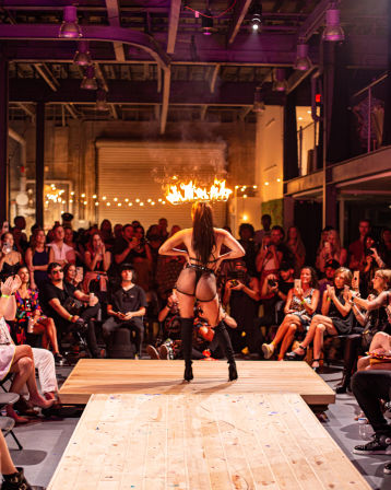 Burlesque-style performer on a wooden runway balancing a flaming crown overhead, wearing fishnet bodysuit and thigh-high boots as a seated crowd watches in an industrial warehouse venue.