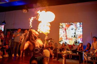 Fire-eater blowing a dramatic flame ball during an indoor art-gallery lounge performance, captivated audience and vibrant pop-art on the wall in the background.