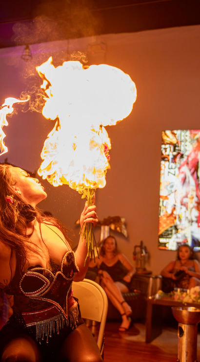 Fire-eater blowing a dramatic flame ball during an indoor art-gallery lounge performance, captivated audience and vibrant pop-art on the wall in the background.
