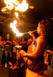 Fire performer in blue lingerie and fishnet stockings sits on a bar counter and breathes a dramatic flame toward the ceiling as an excited nightclub crowd films and cheers.