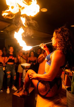 Fire performer in blue lingerie and fishnet stockings sits on a bar counter and breathes a dramatic flame toward the ceiling as an excited nightclub crowd films and cheers.