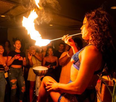 Fire performer in blue lingerie and fishnet stockings sits on a bar counter and breathes a dramatic flame toward the ceiling as an excited nightclub crowd films and cheers.