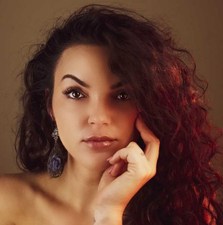 Close-up studio portrait of a woman with long curly dark hair, dramatic brows and glossy lips, ornate dangling earring and hand resting on her cheek in warm, moody lighting.