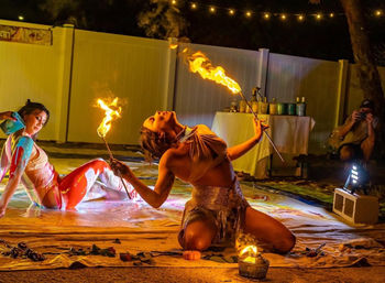 Nighttime backyard fire performance: two artists on a painted canvas floor — one kneeling in a sparkly costume breathing fire from torches, the other reclining with colorful body paint, string lights and onlookers in the background.
