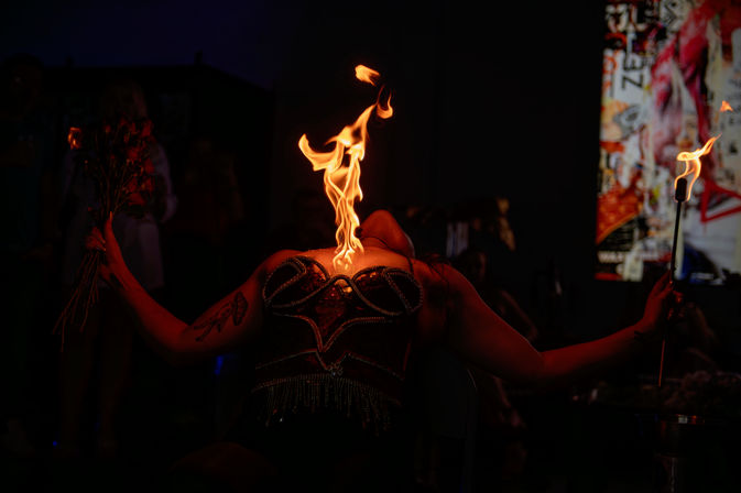 Burlesque fire performer on a dark nightclub stage, reclining in a sequined corset as a tall flame rises from her chest, holding a lit torch and a bouquet with a tattooed arm, blurred audience in background.