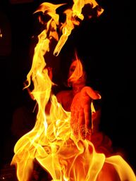 Close-up of a fire performer extending an outstretched hand into swirling orange-yellow flames against a dark background, dramatic fire‑dancing scene.