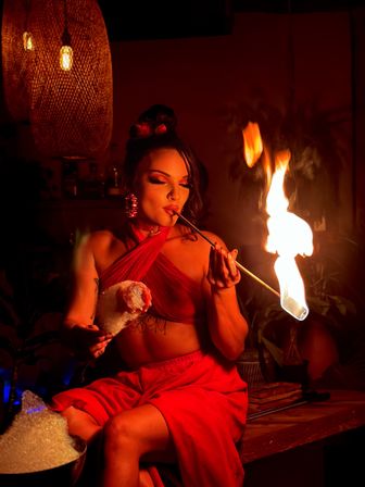 Fire performer in a red outfit at a dim indoor lounge, blowing on a flaming torch held on a metal skewer above a sugar-coated dessert under warm pendant lighting.
