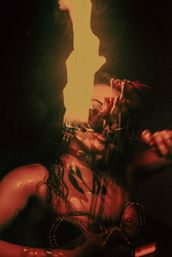 Close-up night portrait of a fire performer holding a tall flame to their mouth beside wilted roses, bathed in red light and ornate jewelry