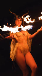 Fire performer in a horned headdress and sheer lace bodysuit twirling flaming wicks in a dramatic nighttime show
