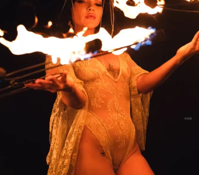 Fire performer in a horned headdress and sheer lace bodysuit twirling flaming wicks in a dramatic nighttime show