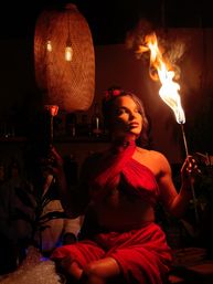Performer in a red wrap top and skirt holding a flaming torch and a rose in a dim, warm-lit bar lounge with woven pendant lights — dramatic nightlife scene