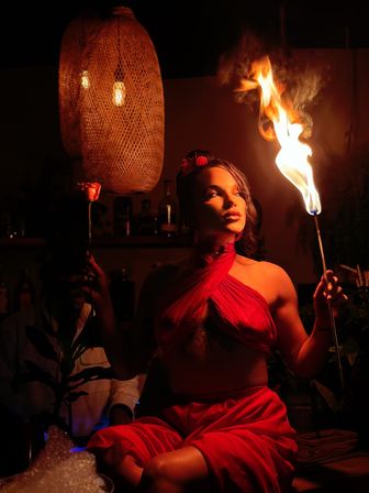 Performer in a red wrap top and skirt holding a flaming torch and a rose in a dim, warm-lit bar lounge with woven pendant lights — dramatic nightlife scene