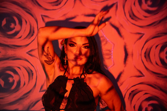 Studio portrait of a woman bathed in red-orange light with rose-pattern wall projection, raised arm casting shadow, visible arm tattoo and cross earring, wearing a black halter top