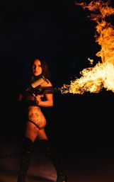 Tattooed woman in lingerie and thigh-high boots firing a flamethrower, dramatic orange flames lighting her against a dark night background