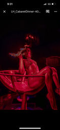 Burlesque performer reclining in a giant martini glass on a red-lit cabaret stage, wearing metallic chains and holding a flaming prop for a dramatic nightclub act
