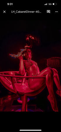 Burlesque performer reclining in a giant martini glass on a red-lit cabaret stage, wearing metallic chains and holding a flaming prop for a dramatic nightclub act