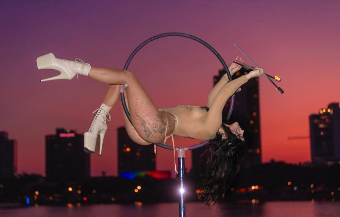 Aerial hoop performer in cream platform heels and fishnet stockings arches back on a suspended hoop while twirling lit fire torches against a pink-orange sunset over a city waterfront skyline.