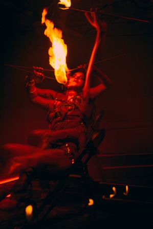 Red-lit fire performer seated on stage, fire-breathing a tall torch flame upward in a dramatic cabaret-style scene