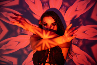 Artistic portrait of a woman with crossed arms against a red floral kaleidoscope projection, dramatic eye makeup and starburst shadow patterns across her face and arms.