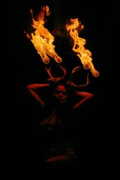 Silhouette of a performer at night holding antler-like props crowned with twin roaring flames, dramatic orange glow against a black background