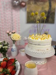 Birthday dessert table with a white semi-naked lemon cake topped with orange slices and yellow billy-ball flowers on a white ceramic stand, surrounded by fresh strawberries, roses and small daisies on a pink tablecloth.