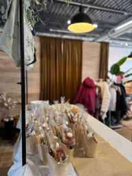 Wrapped mini charcuterie cones displayed on a table at an indoor pop-up market or boutique, with hanging coats, wood-slat wall and warm pendant lighting.