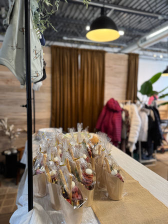 Wrapped mini charcuterie cones displayed on a table at an indoor pop-up market or boutique, with hanging coats, wood-slat wall and warm pendant lighting.