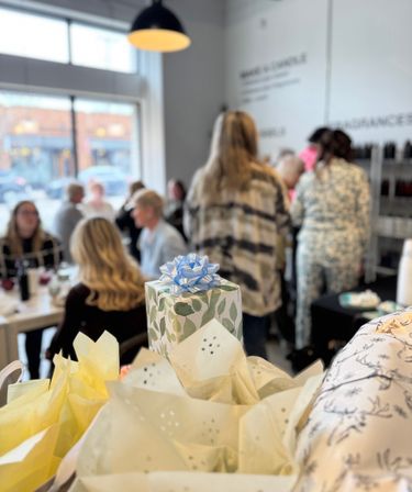 Leaf-pattern gift topped with a blue bow and tissue paper in the foreground, with a blurred group of people chatting inside a bright downtown shop or cafe by a large window and hanging light — casual celebration scene.
