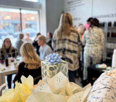 Leaf-pattern gift topped with a blue bow and tissue paper in the foreground, with a blurred group of people chatting inside a bright downtown shop or cafe by a large window and hanging light — casual celebration scene.