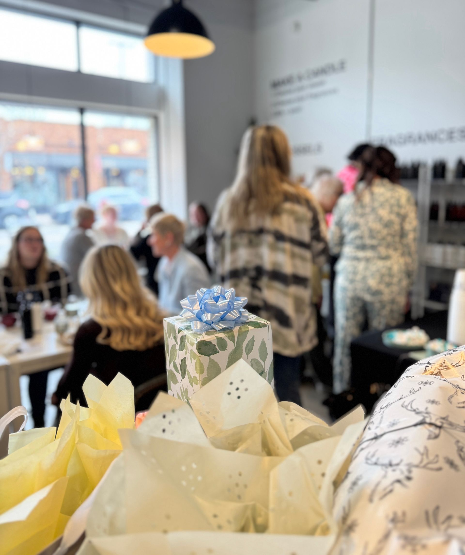 Leaf-pattern gift topped with a blue bow and tissue paper in the foreground, with a blurred group of people chatting inside a bright downtown shop or cafe by a large window and hanging light — casual celebration scene.