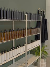 Neat retail shelf display against a muted green wall with rows of black, amber, and white refill squeeze bottles lined up, labeled caps, black aprons on hooks and a potted plant nearby.