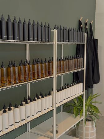 Neat retail shelf display against a muted green wall with rows of black, amber, and white refill squeeze bottles lined up, labeled caps, black aprons on hooks and a potted plant nearby.