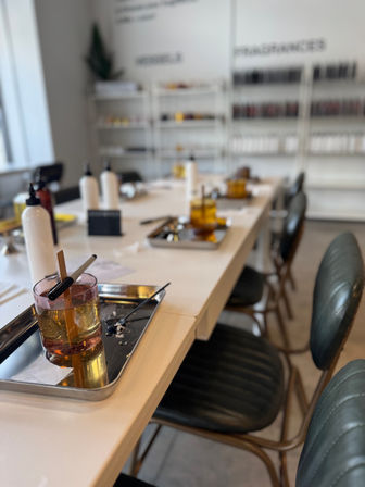 Manicure studio table set for nail dipping with metal trays, amber dipping bowls, wooden sticks, white product bottles, dark green chairs and blurred shelves of nail polish in the background.