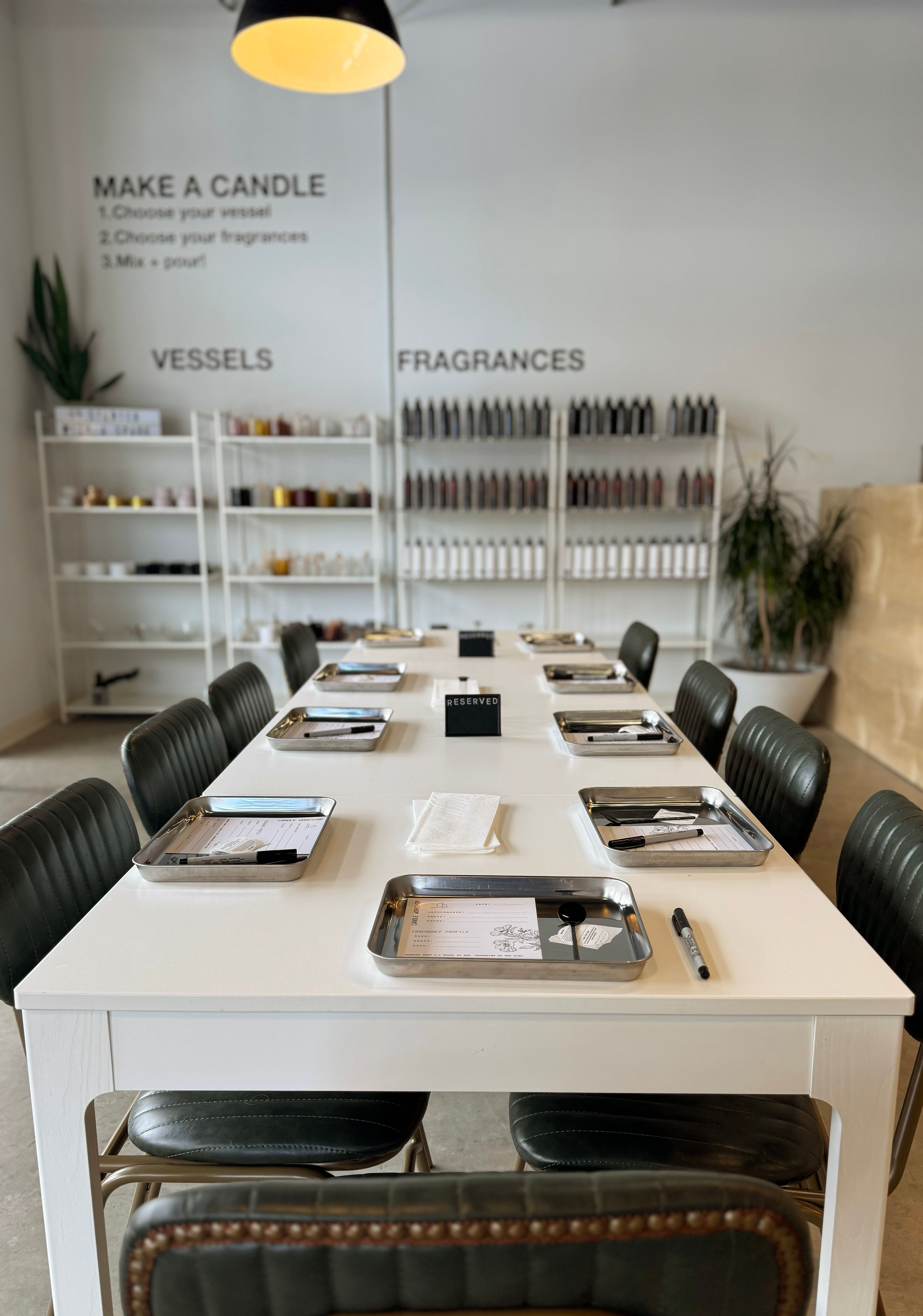 Bright modern candle-making workshop with a long white communal table set with metal trays, spoons and pens, a small ‘reserved’ sign, and shelves of vessels and fragrance bottles in the background.