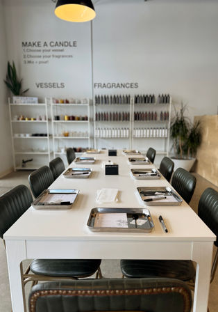 Bright modern candle-making workshop with a long white communal table set with metal trays, spoons and pens, a small ‘reserved’ sign, and shelves of vessels and fragrance bottles in the background.