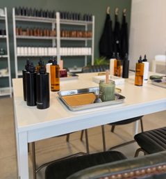 Bright beauty salon waxing station: jar of green wax with wooden spatula on a metal tray, amber and black product bottles on a white treatment table, shelves of skincare bottles and hanging aprons against a green wall in the background.
