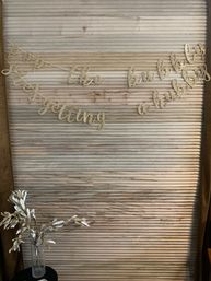 Gold glitter cursive banner with a celebratory phrase draped across a rustic pale wooden slat backdrop above a vase of white decorative branches — indoor bridal shower or engagement party backdrop.