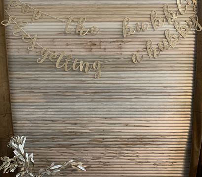 Gold glitter cursive banner with a celebratory phrase draped across a rustic pale wooden slat backdrop above a vase of white decorative branches — indoor bridal shower or engagement party backdrop.
