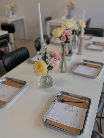 Bright studio table set for a candle workshop with metal trays holding forms and spoons, small vases of yellow and pink roses, and white taper candles