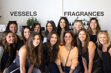 Smiling group of women posing inside a boutique fragrance studio in front of 'Vessels' and 'Fragrances' signage, with shelves of bottles and potted plants.