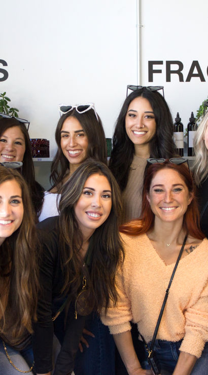 Smiling group of women posing inside a boutique fragrance studio in front of 'Vessels' and 'Fragrances' signage, with shelves of bottles and potted plants.